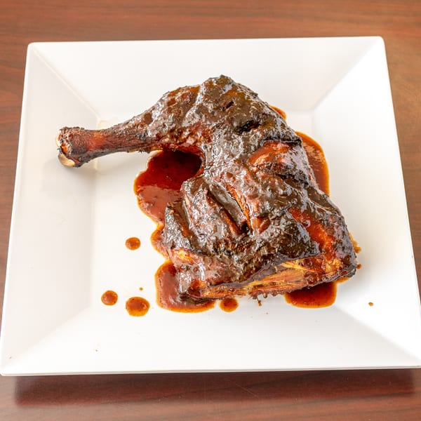 A large piece of dark, glazed jerk chicken with sauce on a white square plate.