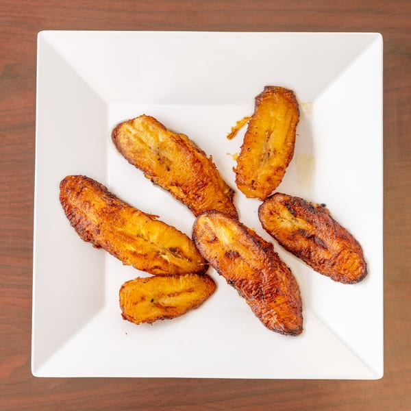 Six golden-brown fried plantains are arranged on a white square plate, on a wooden table.