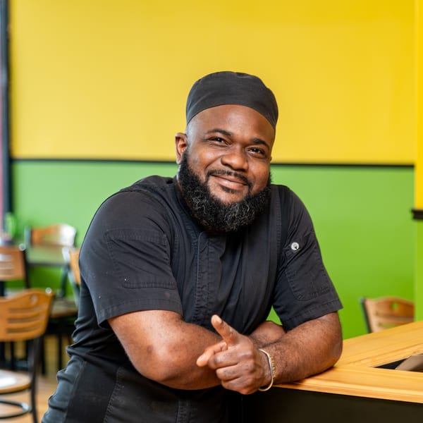 Smiling chef in black uniform and cap leaning on a wooden counter, pointing forward.